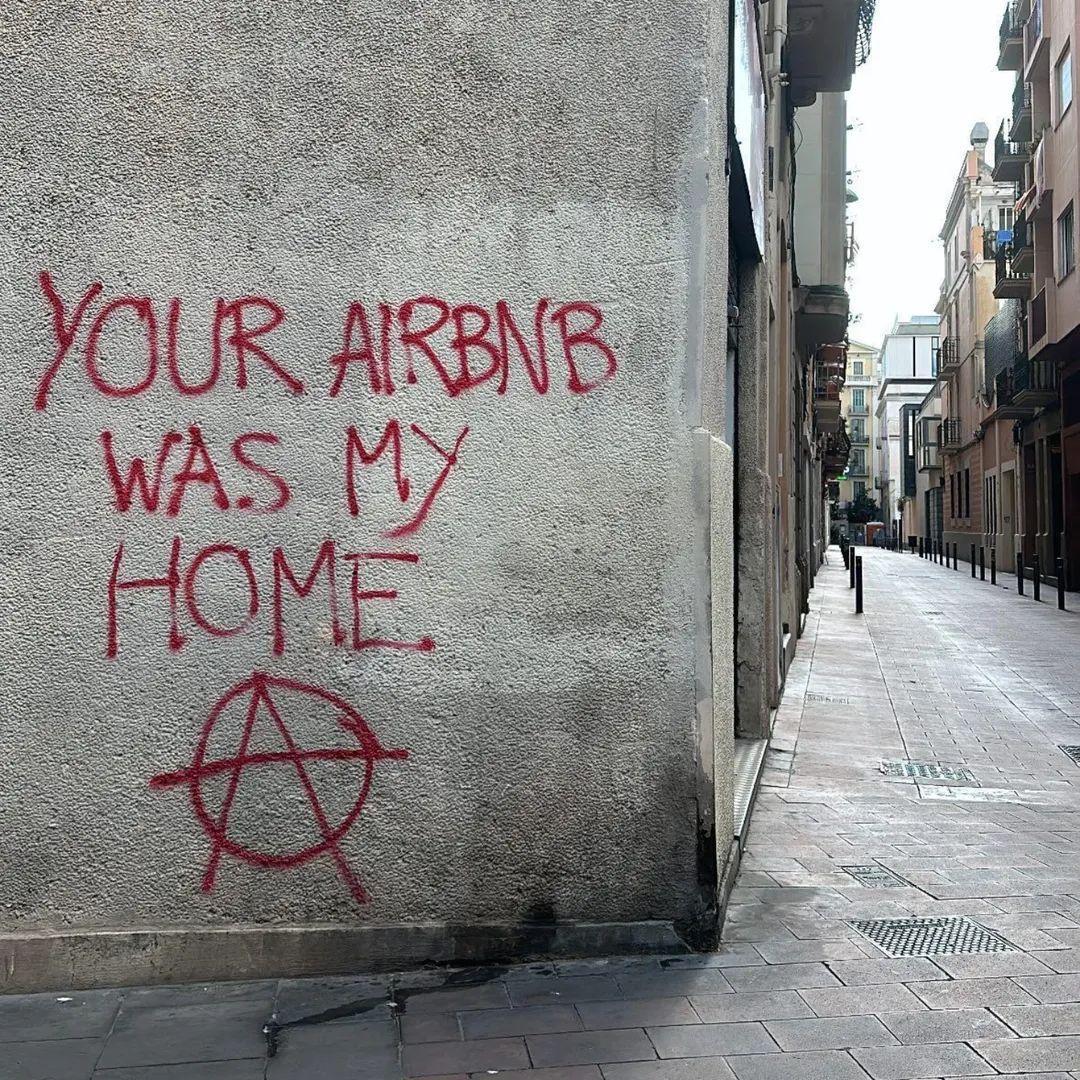 Un muro binaco di Barcellona con una scritta spray rossa: "YOUR AIRBNB WAS MY HOME" e la (A) simbolo dell'anarchia.